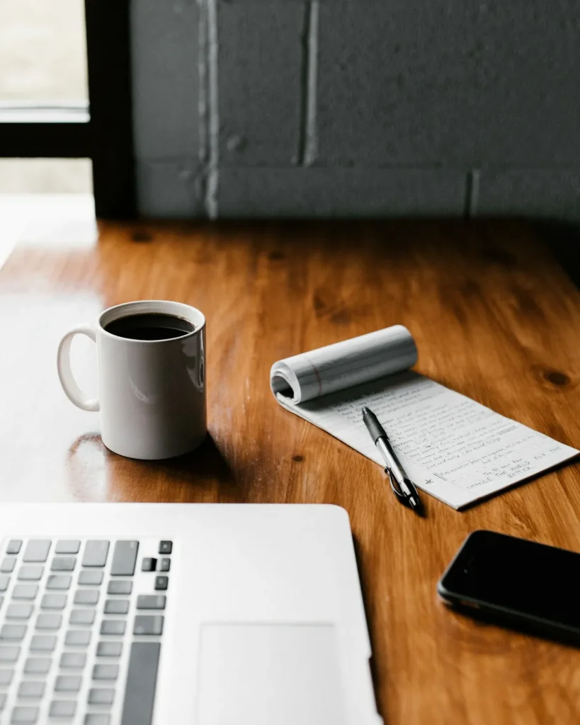 A notebook, pen, phone and coffee beside a laptop on a wooden desk