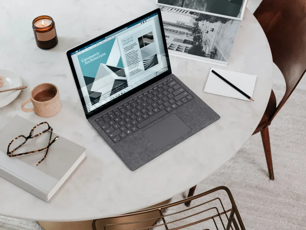 A Surface laptop on a round marble table with a notebook and glasses